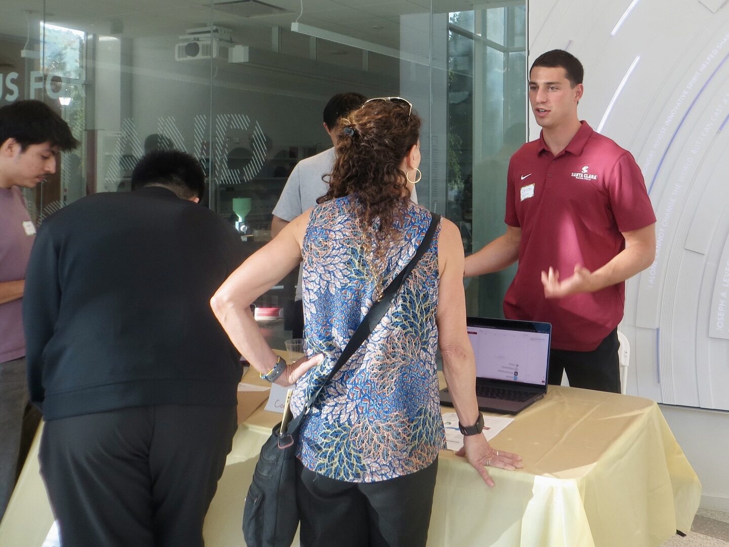 A presenter in an SCU polo explains his project to a visitor at a demo table.